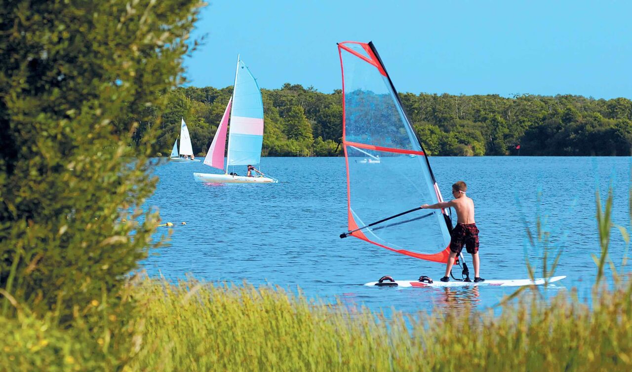 Young windsurfer and sailboats on the lake at LIBRANOO Naturiste Arnaoutchot campsite, Landes, France.