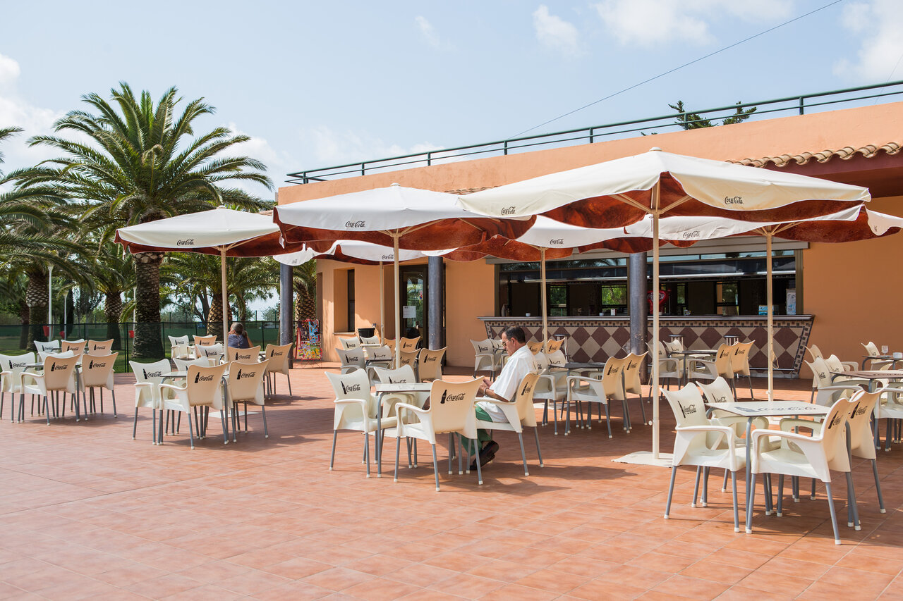 Au�enterrasse des Restaurant-Bars mit Tischen, Sonnenschirmen und Palmen auf dem Campingplatz CAPFUN Aube in Deltebre, Tarragona (43).