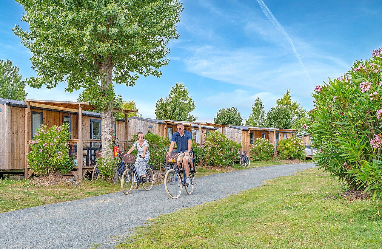 Paar auf Fahrr�dern vor Holz-Mobilheimen auf dem Campingplatz CLICOCHIC Brandais.