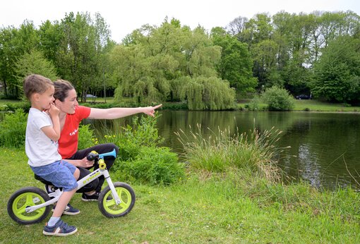 Kind auf Laufrad und Frau zeigt auf den See auf Camping CAPFUN Breteche in Les Epesses (85).