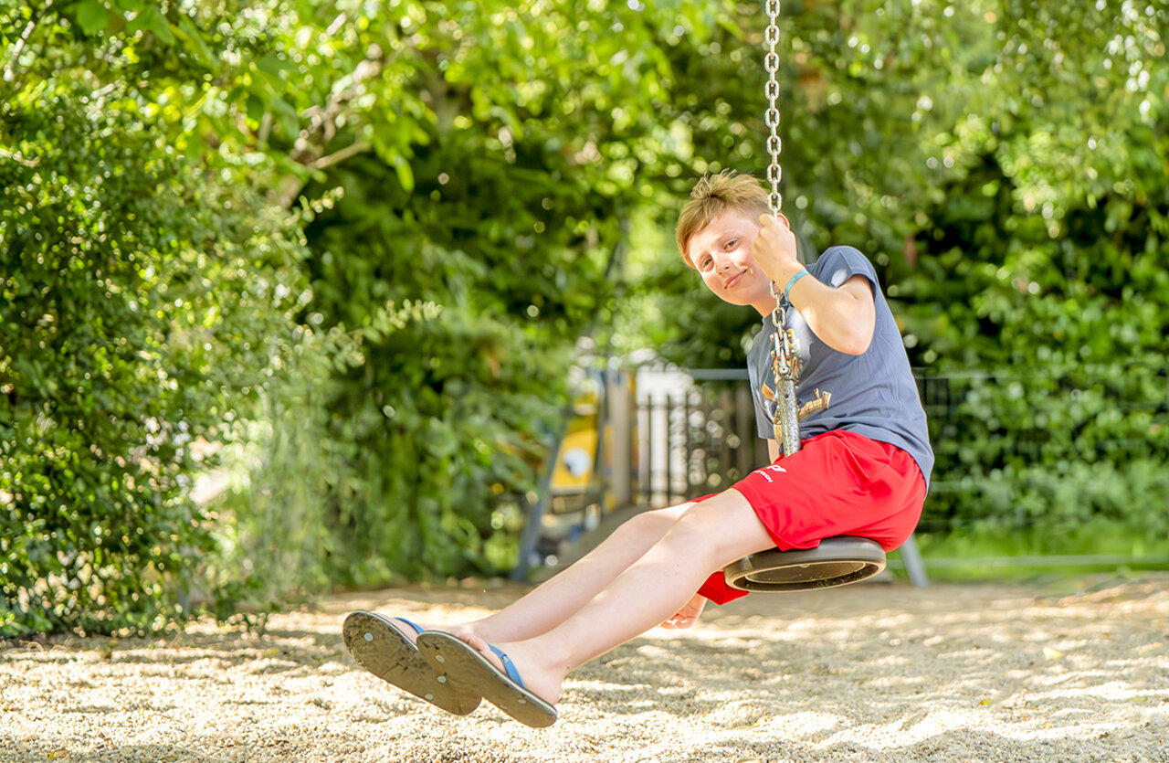 Smiling child on a swing in the playground at VAGUES OCEANES Chataigneraie campsite in Prats-de-Carlux (24).