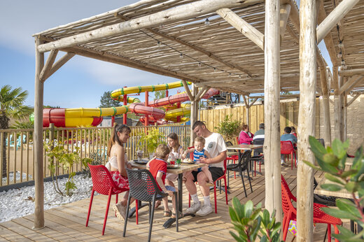 Famille déjeunant en terrasse du restaurant, toboggans aquatiques au camping CAPFUN Chateau d'Arvid (14).