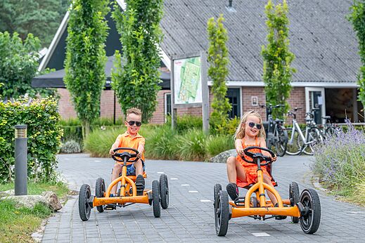 Lächelnde Kinder auf orangefarbenen Tret-Karts auf dem Campingplatz CAPFUN De Belten in Rheeze.