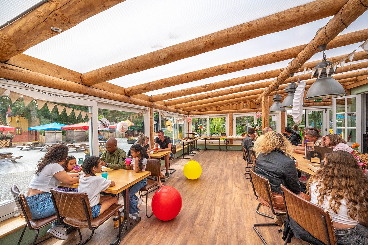 Familien und Kinder speisen im �berdachten Restaurant mit Blick auf den Spielplatz auf dem Campingplatz CAPFUN De Scheepsbel in Doornspijk.