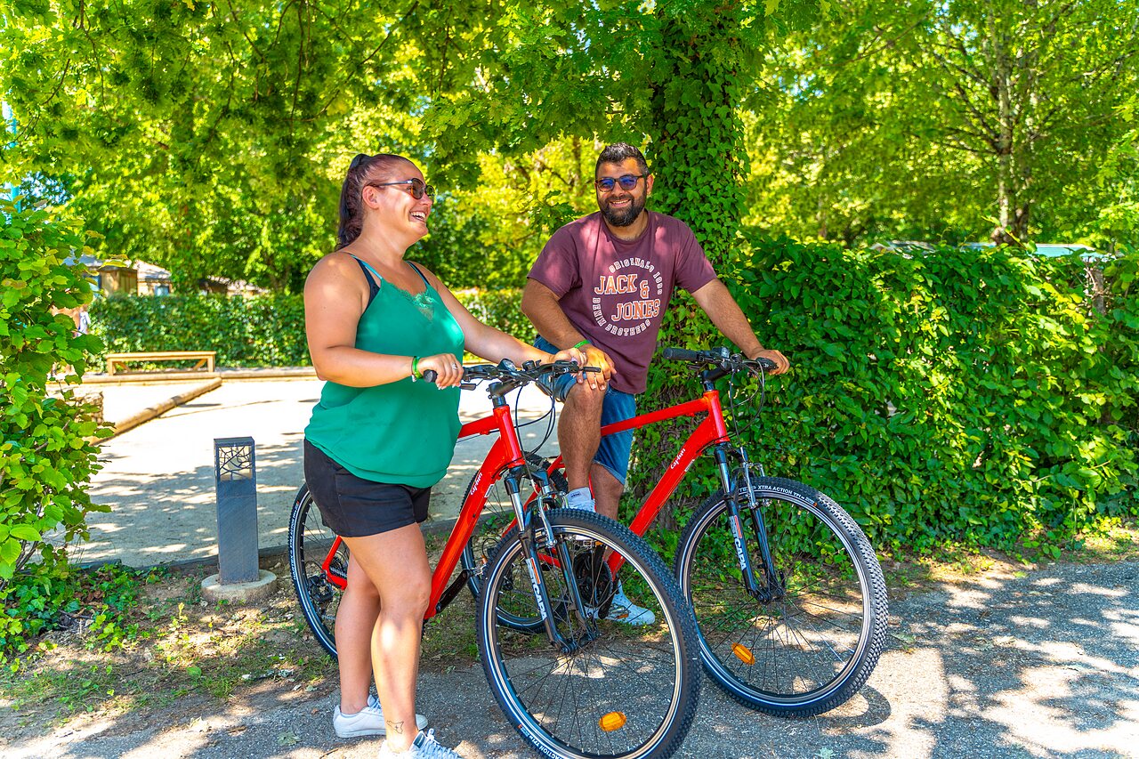 Pareja sonriente con bicicletas rojas, rodeada de naturaleza, en el camping CAPFUN Duravel en Duravel (46).