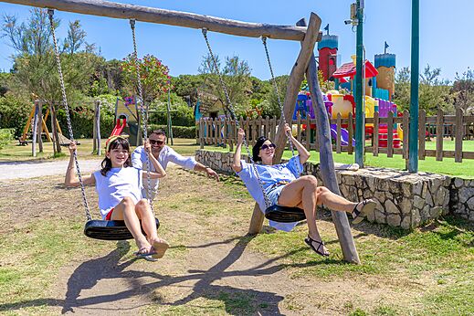 L�chelnde Familie auf den Schaukeln des Spielplatzes auf dem Campingplatz CAPFUN Europing in Tarquinia (01).