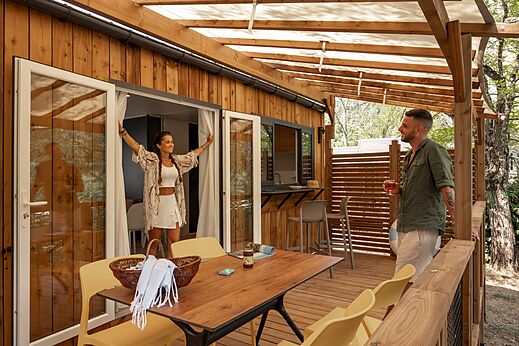 Modernes Mobilheim mit großer Holzterrasse und lächelndem Paar auf dem Campingplatz CLICOCHIC Eyrieux in Les OLLIERES-SUR-EYRIEUX.