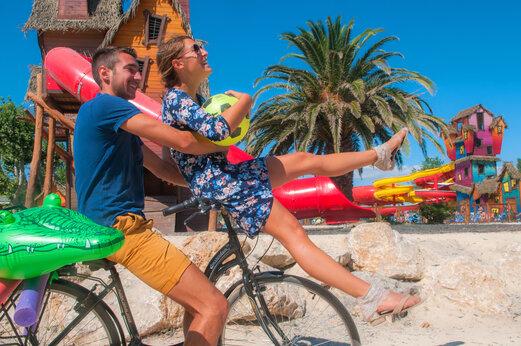 Fr�hliches Paar auf Fahrrad mit Schwimmreifen, bunte Wasserrutschen auf Camping CAPFUN F�erix in MARSEILLAN PLAGE.