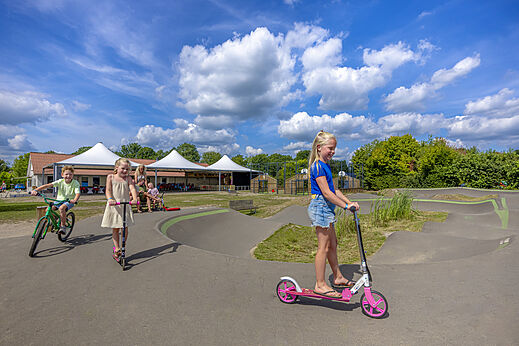 Kinder spielen auf dem Pumptrack mit Rollern und Fahrrad auf dem Campingplatz CAPFUN De Fruithof in Klijndijk.