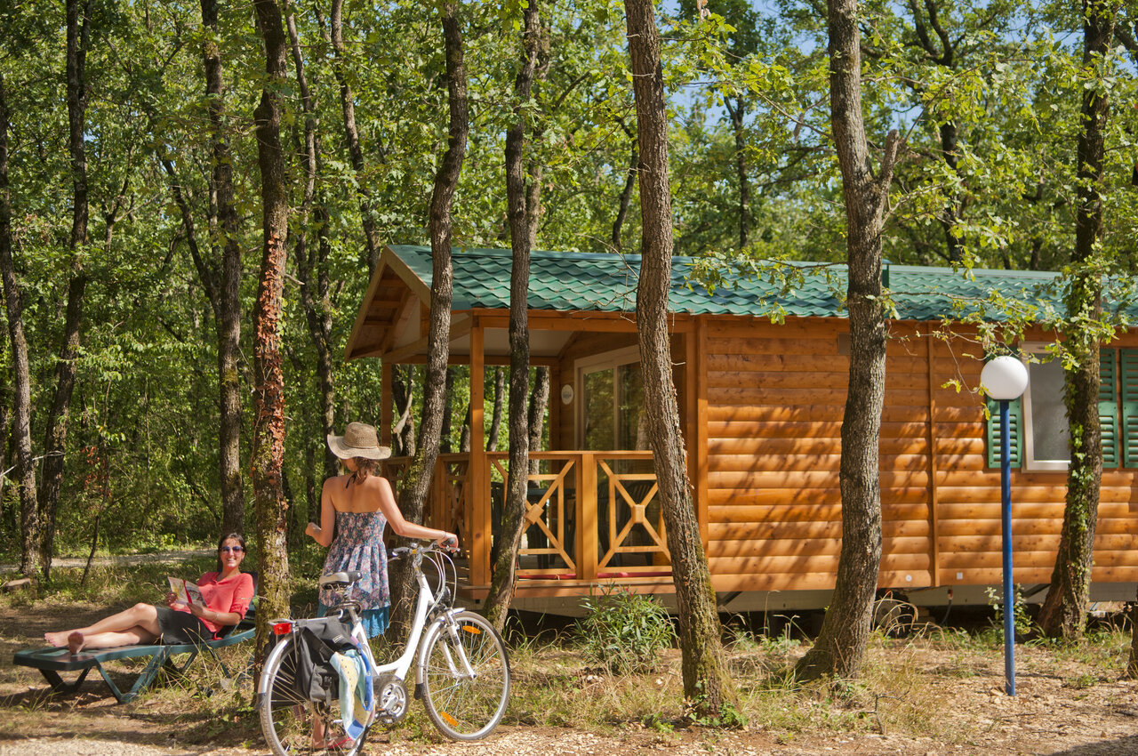 Chalet en bois, femme � v�lo et autre lisant, sous les arbres au camping CAPFUN Grand Lierne � CHATEAUDOUBLE (26).