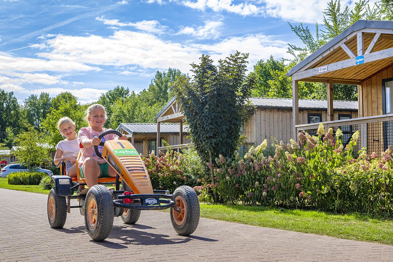 Lachende Kinder auf Tretkart vor Chalets auf dem Campingplatz CAPFUN IJsselstrand in Doesburg.