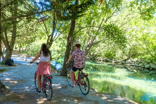 Paar fährt Fahrrad entlang eines schattigen Flusses auf dem Campingplatz CAPFUN Jantou in LE THOR (84).