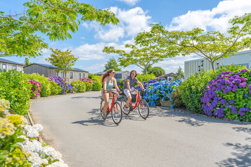 Zwei Personen fahren Fahrrad auf blumiger Allee auf Camping CAPFUN Kervel in PLONEVEZ PORZAY (29).
