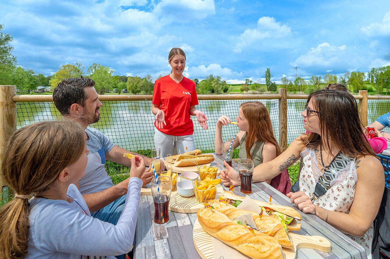 Familie isst auf Terrasse mit Seeblick, auf Campingplatz CAPFUN Mirabelle in VOLSTROFF (57).