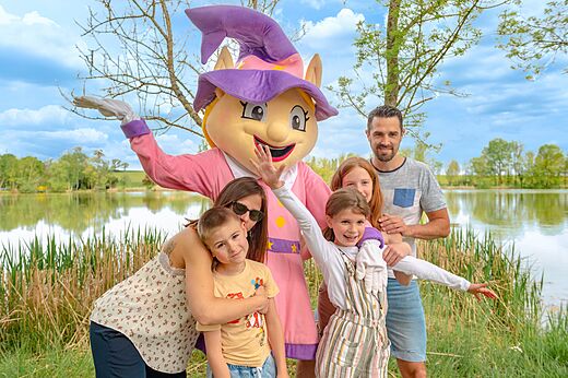 Maskottchen und lächelnde Familie am See auf dem Campingplatz CAPFUN Mirabelle VOLSTROFF.