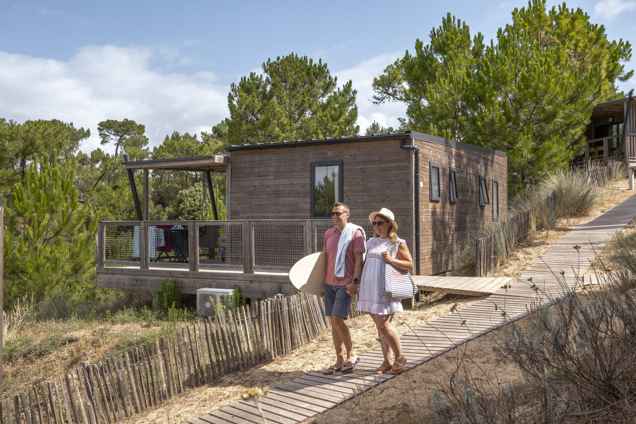 Modernes Holz-Mobil-home mit Terrasse, Paar und Bodyboard auf dem Campingplatz CLICOCHIC Petit Bec in St Hilaire de Riez (85).