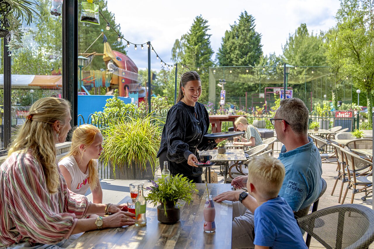 Familie isst auf Terrasse, Kellnerin, Spielplatz auf Camping CAPFUN Ponderosa in Ulicoten.