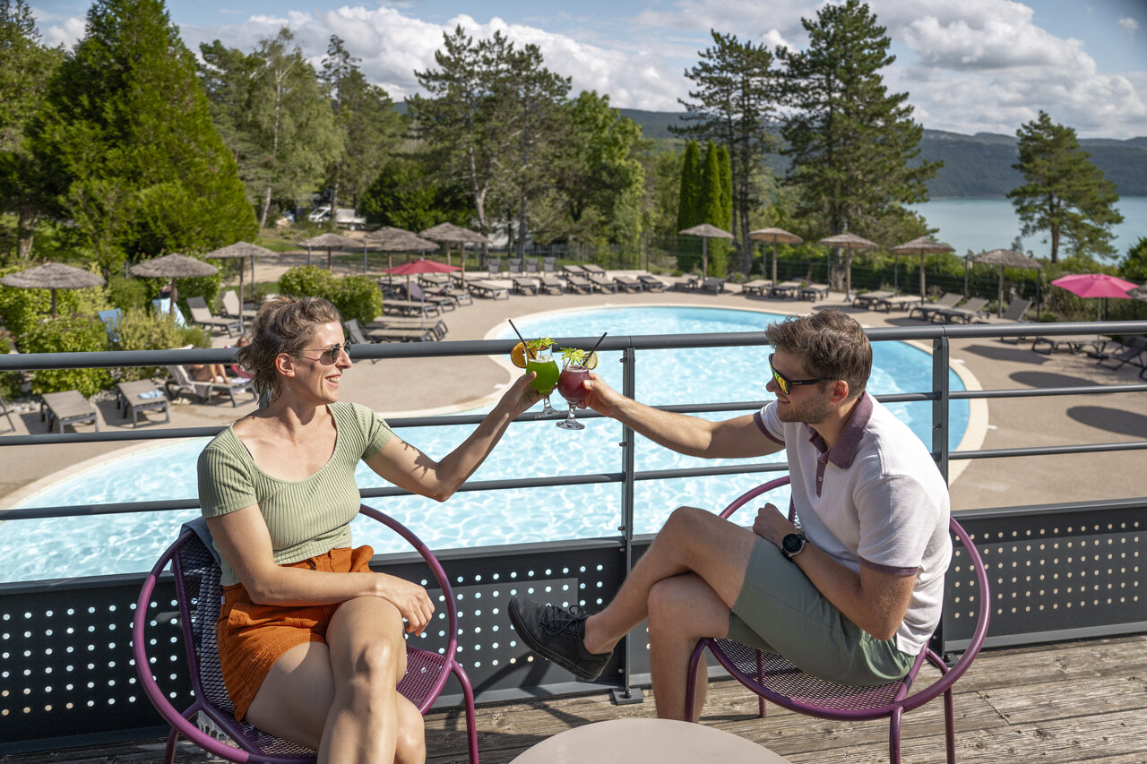 Couple toasting with cocktails, outdoor pool and lake at CLICOCHIC Trelachaume campsite.