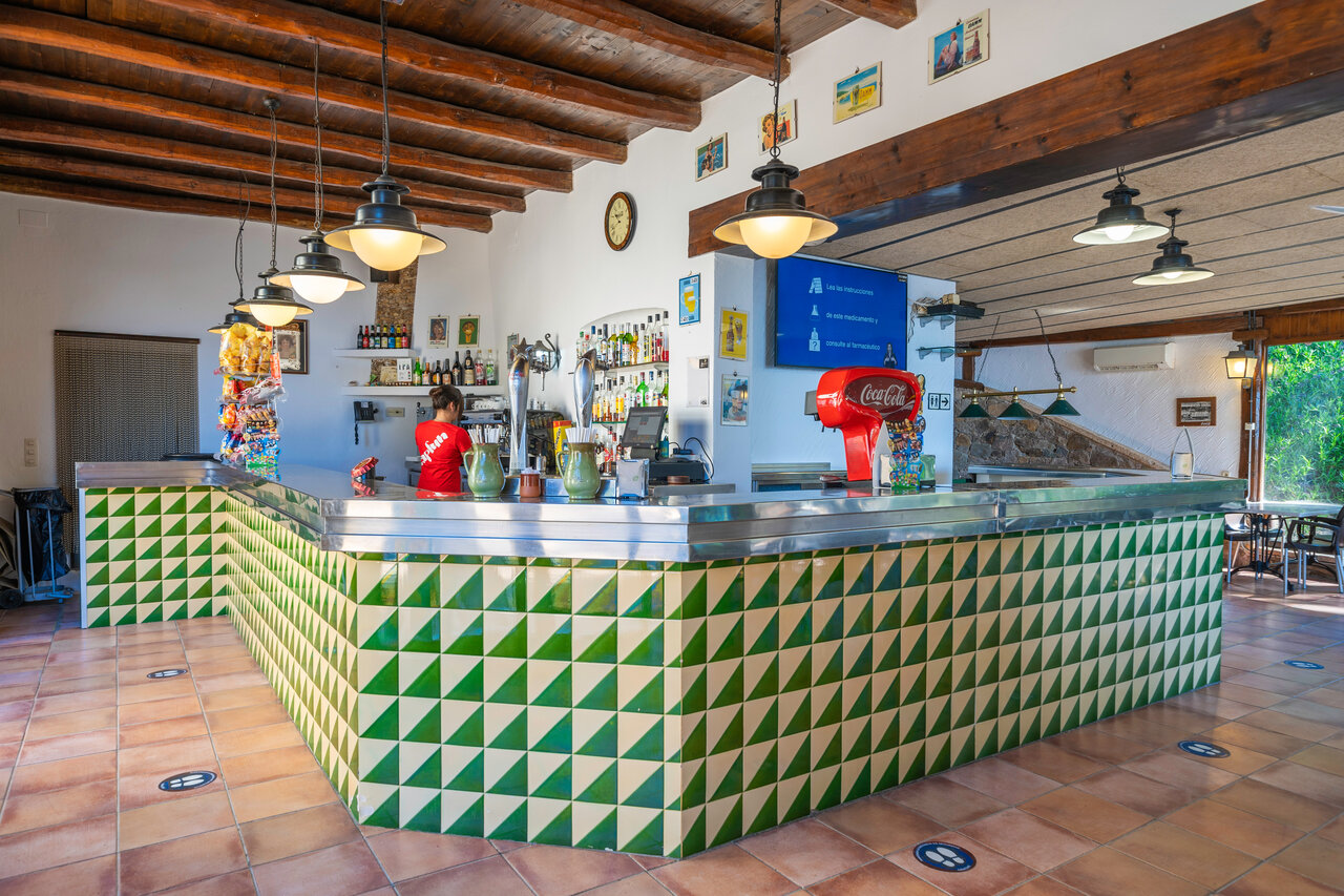 Bar interior con mostrador y camarera en el camping CAPFUN Treumal en Platja d'Aro.